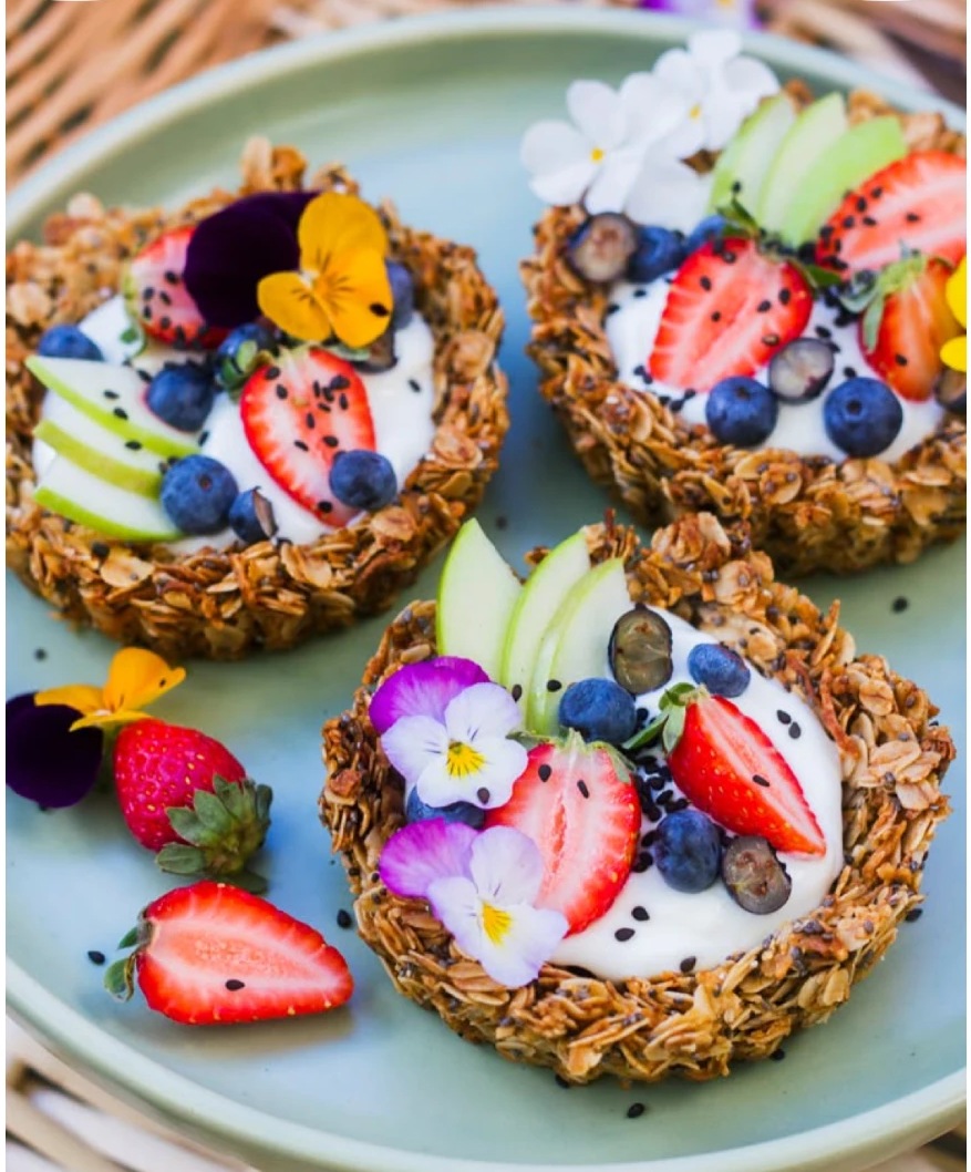 Trois tartes de granola garnies de yaourt, fraises, myrtilles et fleurs comestibles, posées sur une assiette verte