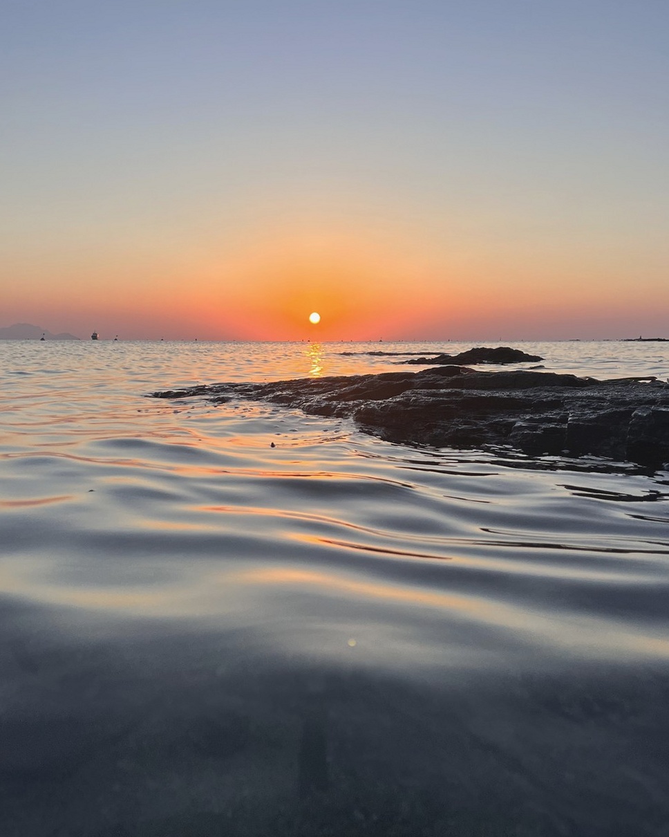 Soleil couchant sur une mer calme, rochers sombres à droite et bateaux à l'horizon