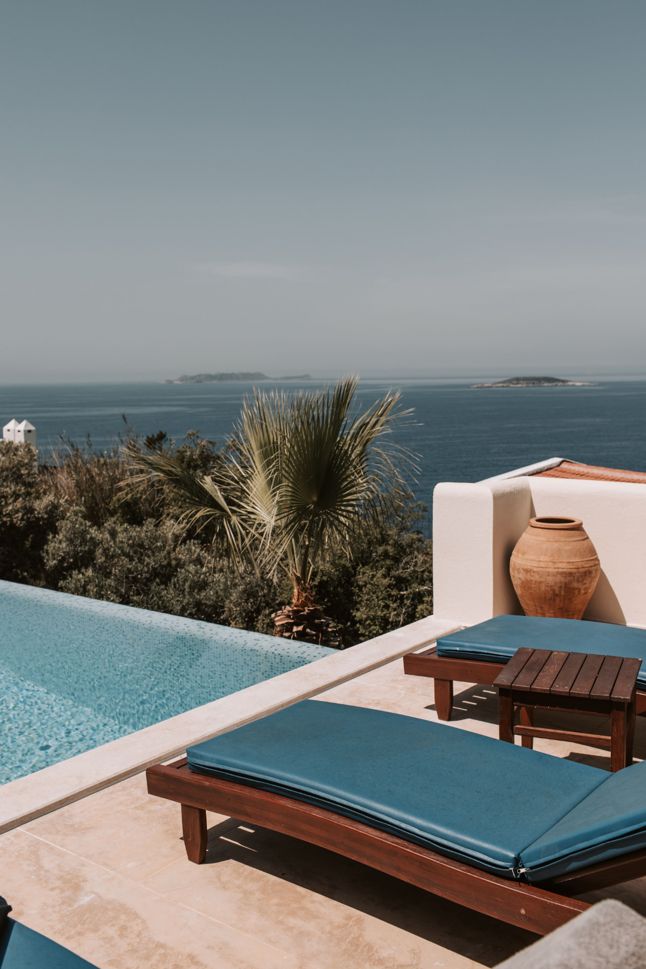 Piscine à débordement face à la mer, transats en bois et coussins bleus sur une terrasse de pierre claire