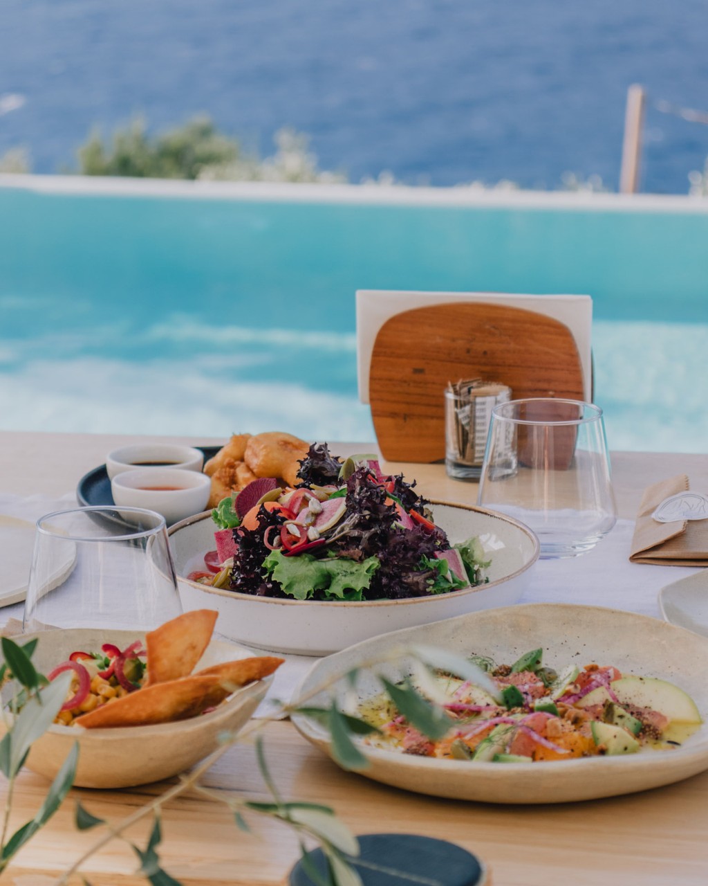 Table dressée au bord d'une piscine à débordement face à la mer : salade, carpaccio et beignets sur céramiques artisanales